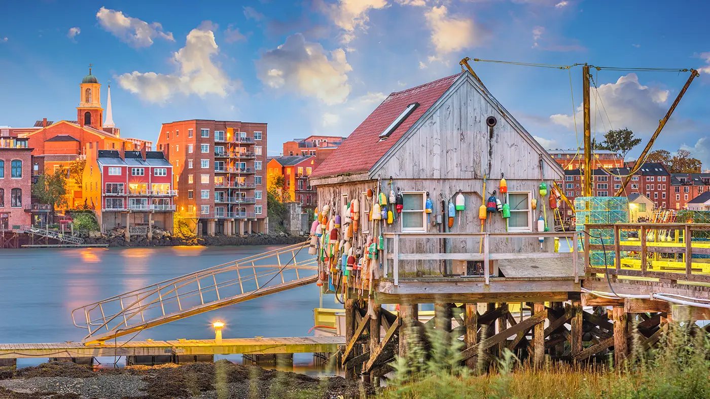 Wooden fishing shack adorned with colorful buoys, set against a backdrop of Southern New Hampshire's waterfront buildings and a vibrant sky, symbolizing local business charm and community support.