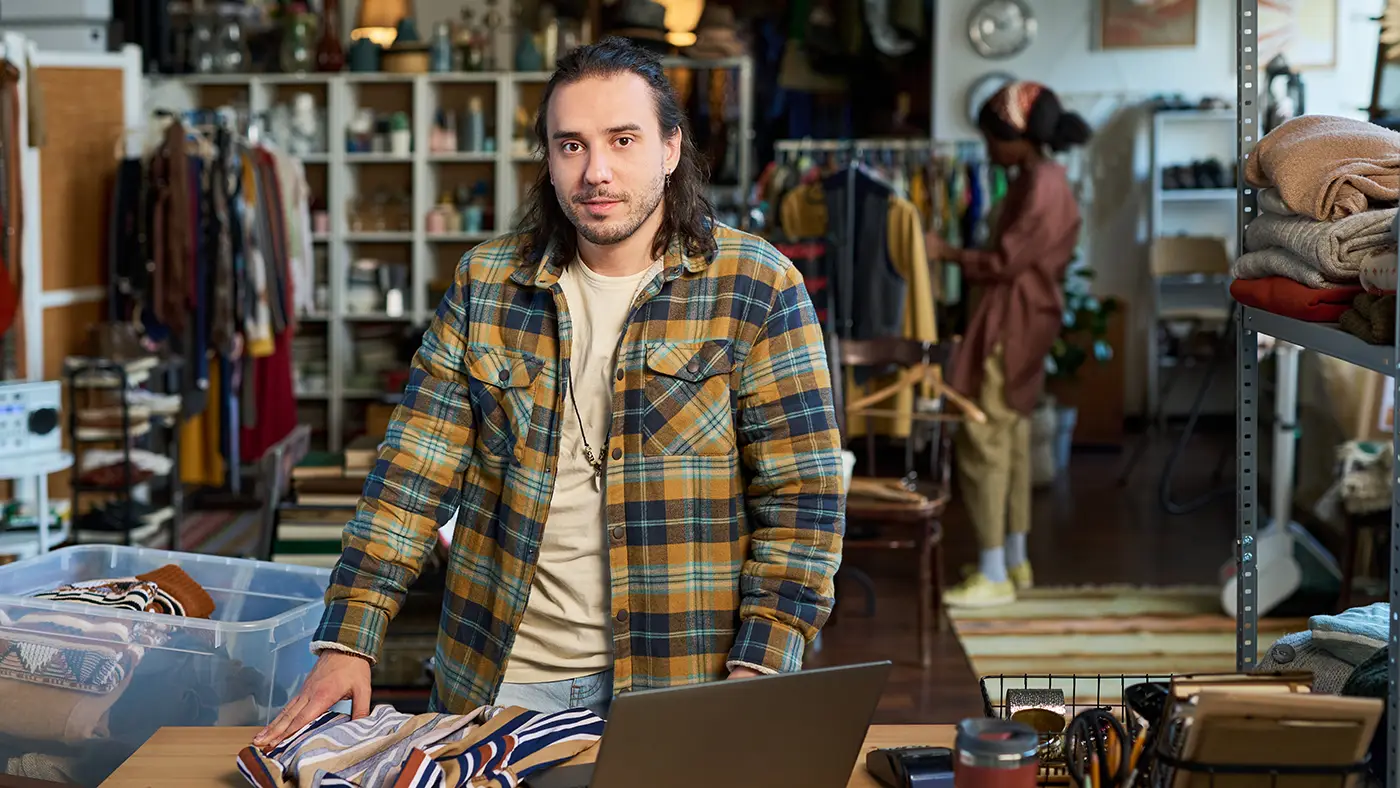 Man in a plaid shirt standing at a table with folded clothes and a laptop, showcasing a local business environment with clothing racks and shelves in a cozy shop setting.